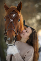 Close portrait of young smiling woman and brown horse at winter sunset. Woman with long hear in sweater near big horse