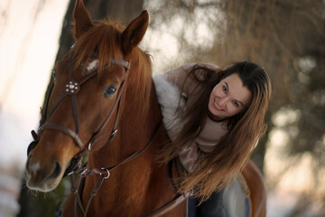 Fototapeta premium Close portrait of young smiling woman with long hair in sweater sitting on brown horse at sunset, winter