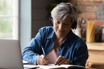 Diligent aged student. Focused elderly hispanic woman in headphones watch webinar on laptop screen...