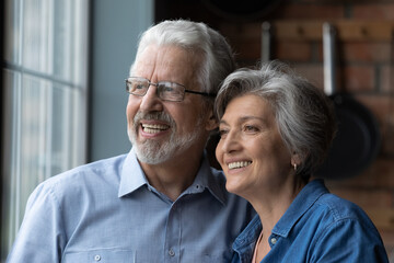 Lost in dreams. Headshot of affectionate elderly married couple pensioners cuddle look away with happy white smiles enjoy love care create plans. Happy healthy aged spouses stand by window think dream
