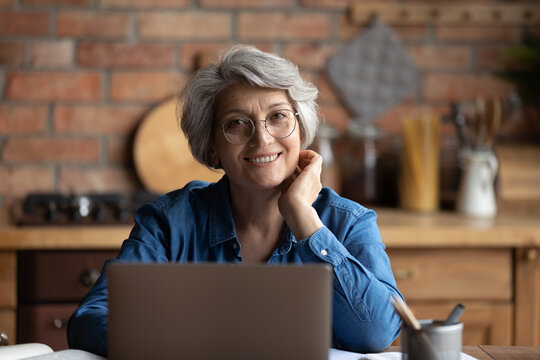 Headshot portrait of happy senior aged hispanic female sit at desk use laptop work study distantly. Confident old age woman remote worker freelancer look at camera at home office workplace at kitchen
