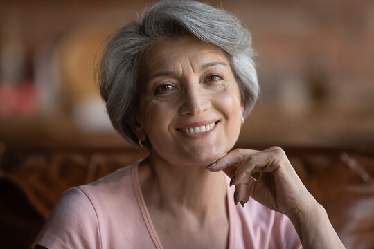 Beautiful age. Headshot portrait of happy mature latin female pensioner looking at camera with white smile. Attractive grey haired 60s retired hispanic woman posing indoors touching chin with finger