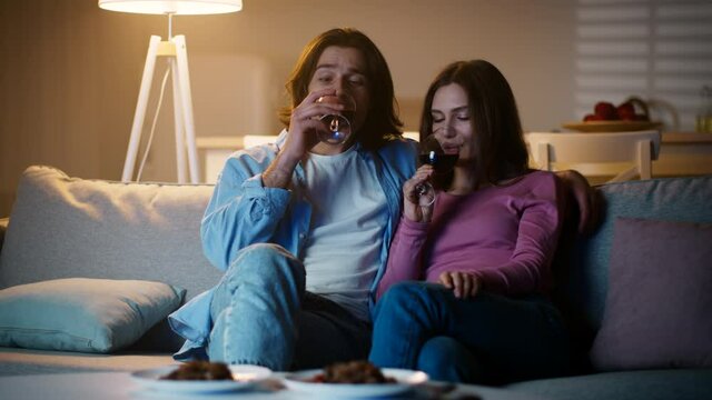 Romantic evening. Young couple in love enjoying red wine and watching tv, sitting on couch at home