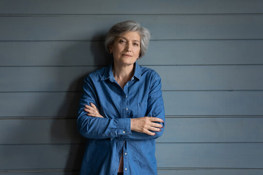 Charismatic person. Serious elderly latin lady posing for portrait keep arms crossed. Confident old female mother grandma in stylish jeans jacket stand by grey planked wall look at camera. Copy space
