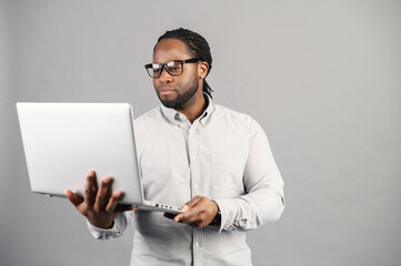 Concept of confident business expert. Serious young handsome black man with elegant glasses in casual shirt, holding laptop, looking at screen, standing against grey background, no time for break