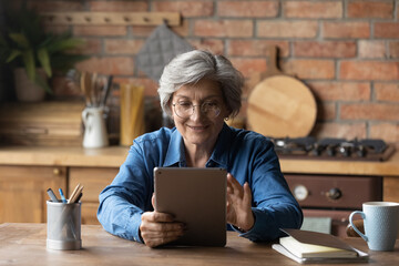 Modern day pensioner. Smiling mature latina woman sit at home office by kitchen table watch video on tab computer screen. Old age grandmother enjoy reading e book on tablet pc scroll news on websites - obrazy, fototapety, plakaty