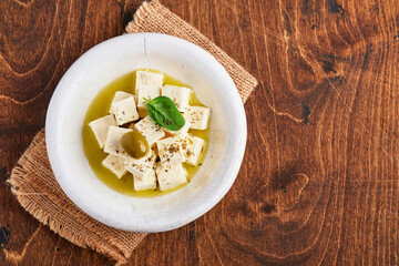 Feta cheese cubes with rosemary, olives and olive oil sauce in white bowl on old brown wooden background. Traditional Greek homemade cheese. Selective focus.