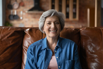 Active old age. Portrait of optimistic retired hispanic female clothed in stylish casual demonstrating perfect white teeth in beautiful smile. Energetic senior latin woman sit on couch look at camera
