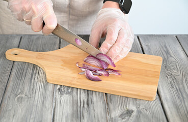 A woman in transparent gloves is cutting an onion with a large kitchen knife on a wooden board.