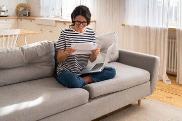 Overjoyed millennial woman sitting on couch at home, holding and reading paper document, feel...