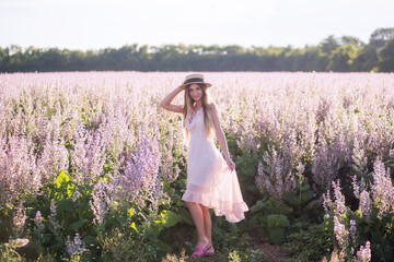 Young blonde woman in light chiffon sundress against the background of blooming field of pink sage. Portrait of beautiful girl holding straw hat. Weekend walk outside the city. Agricultural texture