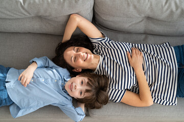 Top above view of happy young mom and little kid son relaxing on sofa at home. Couple of carefree mother and cute child lying, resting on couch, having fun, looking together at camera. Family, love.