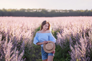 A young woman in a blue shirt and denim shorts stands by a blooming pink sage field. Portrait of girl with long blond hair wearing a straw hat at sunset. Flower texture. Travel out of town. Copy space