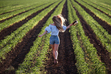 Young woman in blue shirt, denim shorts runs along green plowed field. Long hair develops at sunset, the girl has straw hat in hands. Promote agricultural perspective, the texture of black soil ground