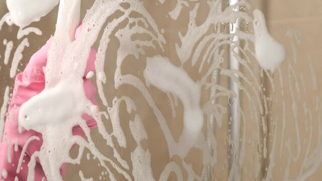 Woman in pink rubber gloves cleans glass door in shower cabin with squeegee and detergent foam. Housewife, housekeeper uses limescale remover for washing in bathroom. Working in home, hotel cleaning 