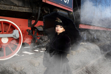 A beautiful girl in a historical retro dress against the background of an old steam locomotive at...
