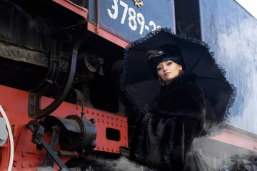 A beautiful girl in a historical retro dress against the background of an old steam locomotive at the station.