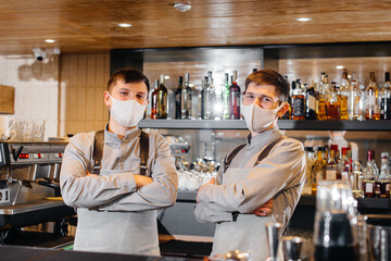 Two stylish bartenders in masks and uniforms during the pandemic, stand behind the bar. The work of restaurants and cafes during the pandemic.