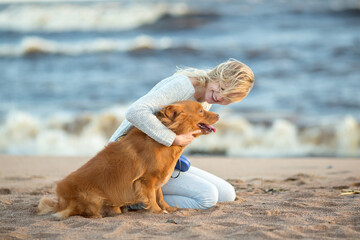 girl in jeans and a blue sweater sitting with his dog on the sand by the sea