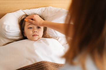 Close-up view of young mother measures temperature touches forehead of sick daughter lying in bed at home. Parent caring for sick child lying in bed with flu. Concept of children health.