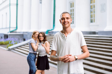 Portrait of a cheerful young boy looking to the camera, two gossiping girls on the background. Don't care what people talk, stay confident.