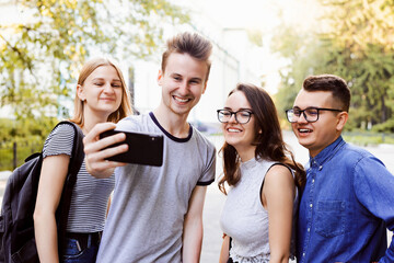 Four friends smiling happily and making selfie on a smartphone. Beautiful summer sunny day, happy students have fun.