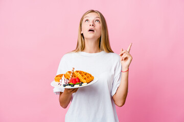 Young russian woman eating a waffle isolated pointing upside with opened mouth.