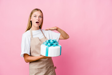 Young russian baker woman holding a delicious cake shocked and amazed holding a copy space between hands.