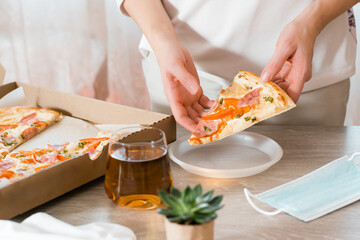Obraz na płótnie Canvas Takeout food. A woman puts a slice of pizza in a disposable plastic plate and a box of pizza on the table in the kitchen.