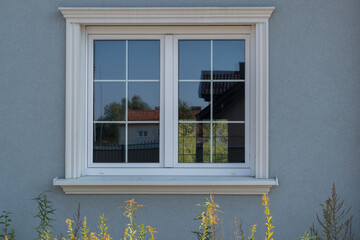 Window with white frame and trim on gray facade of newly built house. Tall grass growing below. On glass can see reflections of houses opposite and contrary window with thickets peeking through it.