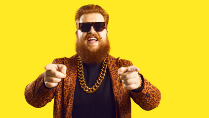 Happy guy in gold necklace and leopard suit pointing fingers at camera isolated on color...