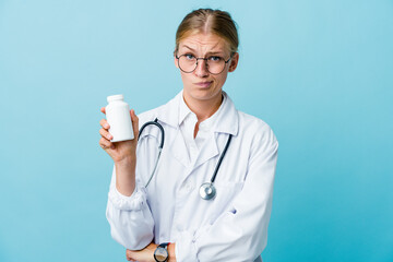 Young russian doctor woman holding pills bottle on blue unhappy looking in camera with sarcastic expression.