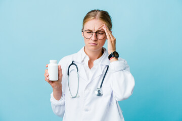 Young russian doctor woman holding pills bottle on blue touching temples and having headache.