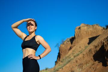 Young athlete woman looking into distance, covering her eyes from the sun, standing on sandy cliff...