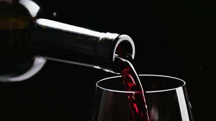 Red wine pouring from bottle into goblet. Close-up of red wine pouring in wine glass at black background. Slow motion. Macro shot
