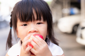 Closeup cute Asian girl is eating apple. Children eat fresh fruit as snack. Child use both hands to control the apples for delicious bite. Asian kids is 3-4 years old.