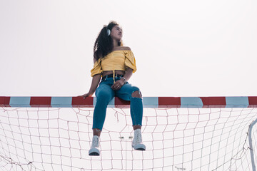 Young African American girl in a yellow t-shirt sitting on a soccer goal listening to music looking at the sky.