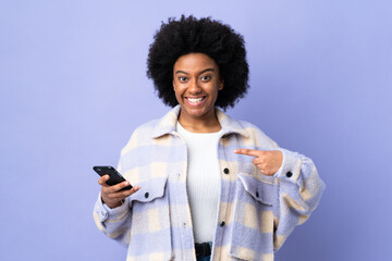 Young African American woman using mobile phone isolated on purple background with surprise facial expression