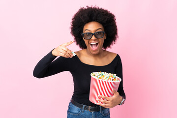 Young African American woman isolated on pink background with 3d glasses and holding a big bucket of popcorns while pointing front