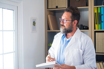 Fototapeta premium Pensive young male caucasian doctor in white medical uniform look in window distance thinking or pondering, serious man GP plan future career or success in medicine, visualize at workplace writing.