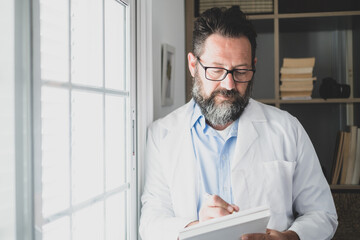 Fototapeta premium Pensive young male caucasian doctor in white medical uniform look in window distance thinking or pondering, serious man GP plan future career or success in medicine, visualize at workplace writing.