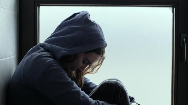 A depressed, sad and lonely young woman with hoody sitting in front of window. Moody and dark atmosphere of Millennial girl at home. Concept of depression, isolation, loneliness, mental health. 4K.