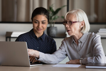 Fototapeta premium Diverse businesspeople sit at table in office look at laptop screen discuss company ideas or project together. Multiracial female colleagues use work online on computer brainstorming cooperating.