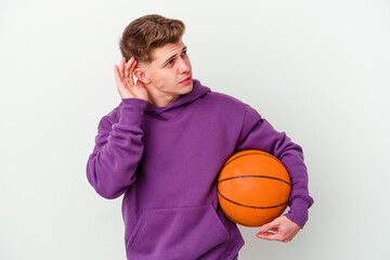 Young caucasian man playing basketball isolated background trying to listening a gossip.