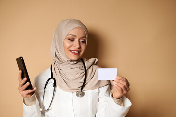 Attractive Arab doctor with covered head in hijab holding a cell phone and a blank plastic card,...