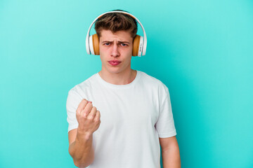 Young caucasian man listening to music with headphones isolated on blue background showing fist to camera, aggressive facial expression.