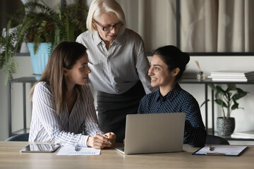 Diverse multiracial female colleagues gather in office work on computer together brainstorm over project startup. Multiethnic women coworkers use laptop talk cooperate at briefing. Teamwork concept.