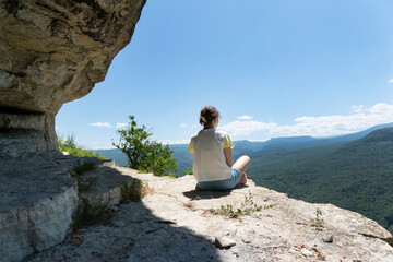Naklejka na ściany i meble Young female sit on rock in high green mountains looking over green forest. Freedom concept, relaxation. Hiking in beautiful places, copy space.