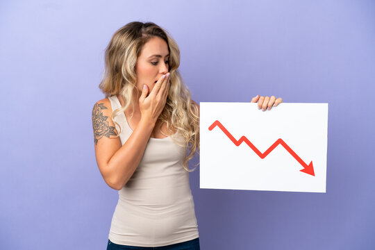Young Brazilian woman isolated on purple background holding a sign with a decreasing statistics arrow symbol with surprised expression