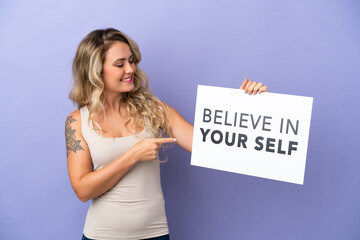 Young Brazilian woman isolated on purple background holding a placard with text Believe In Your Self and pointing it